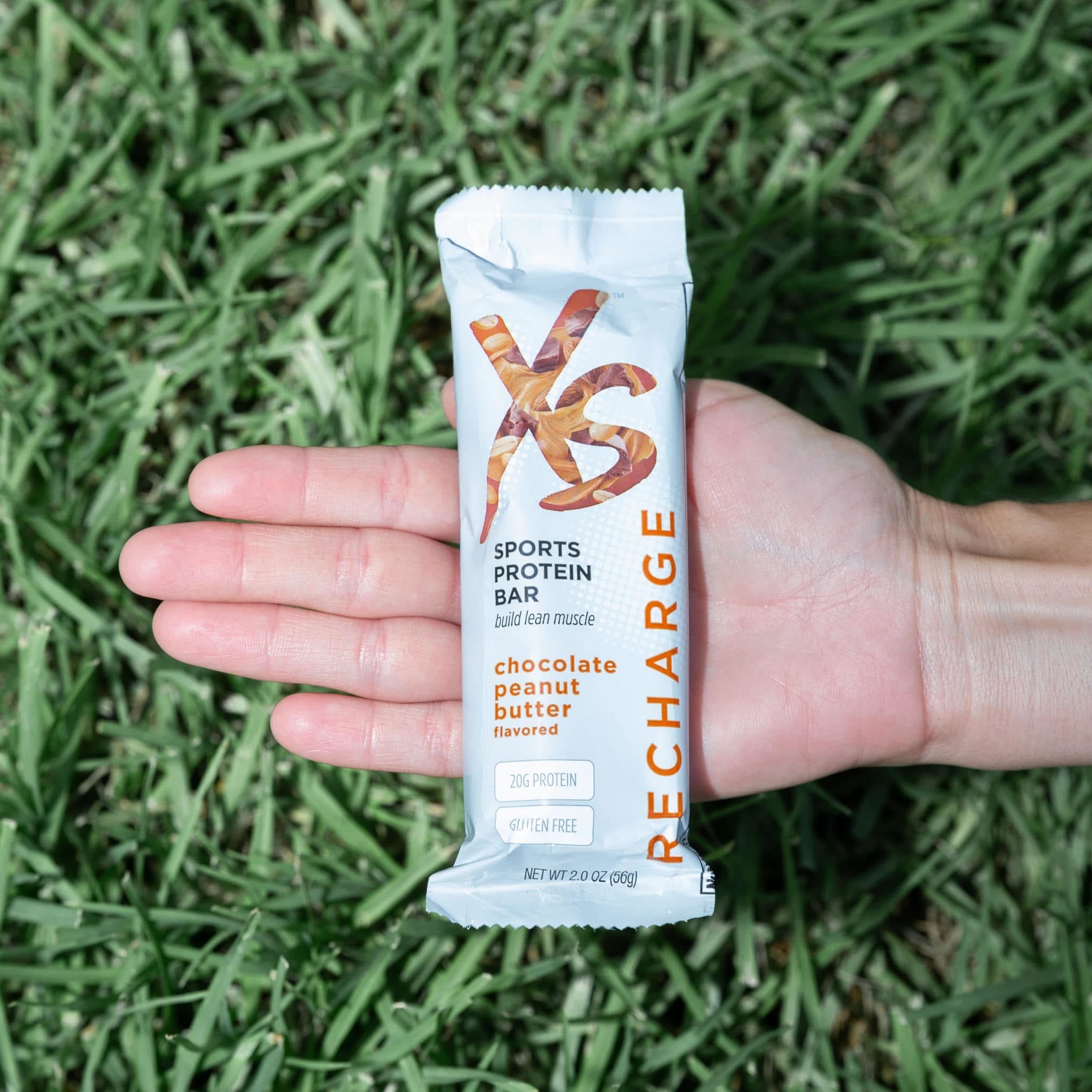A cropped image of a hand outstretched over a patch of lawn is holding an XS sports protein bar. Get ready for what's next - XS Sports Protein Bars help repair and build lean muscle mass and are perfect for between and after workouts.