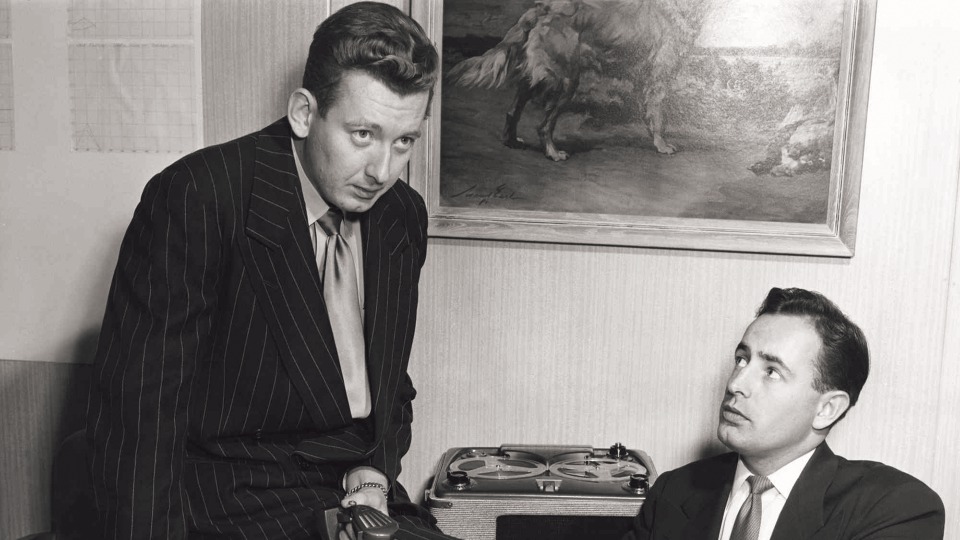 Vintage photo of Rich DeVos and Jay Van Andel in 1952. Jay is sitting on top of the desk while recording a taped message while Rich is seated in a chair next to him.